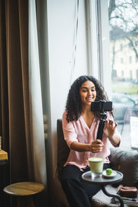 Smiling young female influencer vlogging through mobile phone on tripod sitting by window at creative office