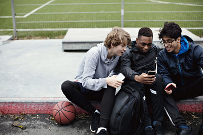 Male friends using mobile phones while sitting on sidewalk after basketball practice during winter