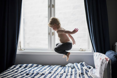 Shirtless boy jumping on bed by window at home