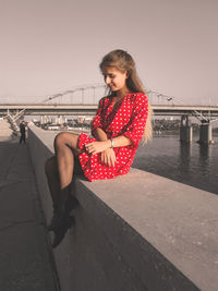 Young woman sitting on railing by bridge against sky