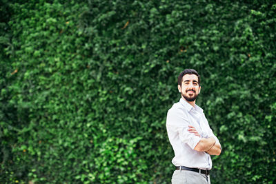 Portrait of young man with arms crossed standing against plants at park