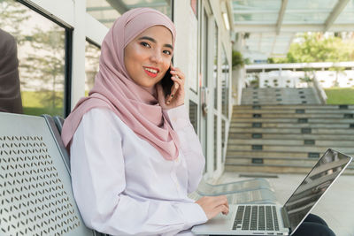 Portrait of smiling young woman talking on mobile phone while sitting with laptop at seat