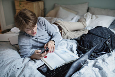 Addictive teenage boy using social media on mobile phone while doing homework in bedroom