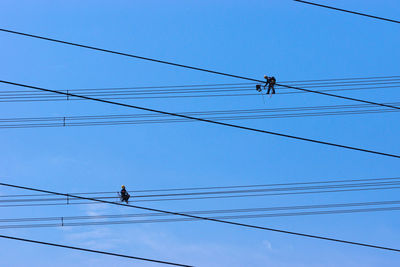 Low angle view of workers working on electricity pylon cables against clear blue sky