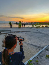 Side view of woman photographing against sky during sunset