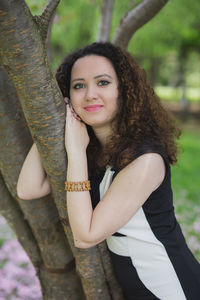 Portrait of smiling young woman leaning on tree trunk in park