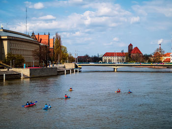 People on river by buildings in city against sky