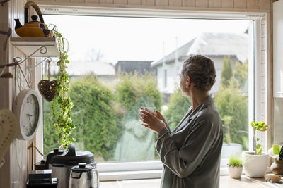 Mature woman holding tea glass at the window in kitchen