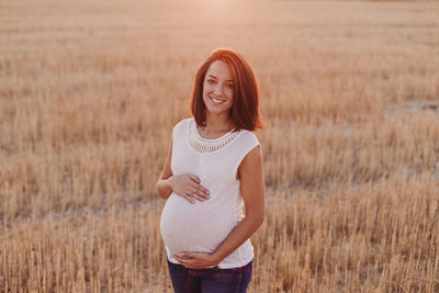 Portrait of smiling pregnant woman with hands on stomach standing on field