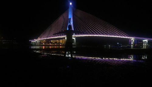 Illuminated suspension bridge over river against sky at night
