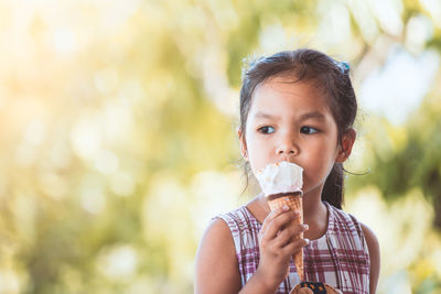 Close-up of cute girl eating ice cream cone while sitting against trees at park