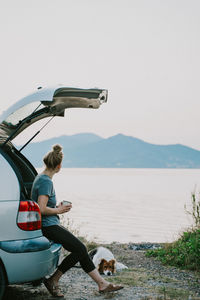 Rear view of woman with dog sitting on shore against sky