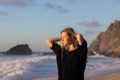 Teenage girl with hands behind head standing at beach against sky during sunset