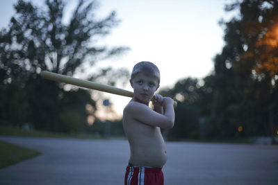 Shirtless boy holding baseball bat against trees at park