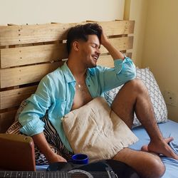 Full length of smiling man sitting with hand in hair on bed at home