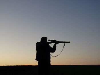 Silhouette of man holding sniper against sky during sunset