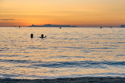 Silhouette people swimming in sea against orange sky