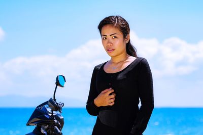 Portrait of young woman standing by motor scooter against sky