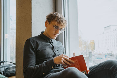 Young blond man reading book by window in university cafeteria