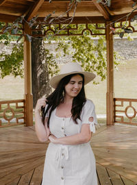 Portrait of beautiful young woman wearing white dress standing under wooden pavilion in park.
