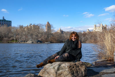 Smiling woman looking away while sitting on rock by lake against sky