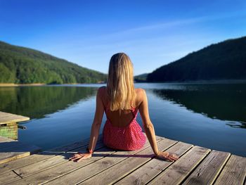 Rear view of blonde woman sitting down on a wooden pontoon and looking at the lake in summer