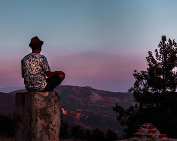 Rear view of man sitting on rock against mountain and sky