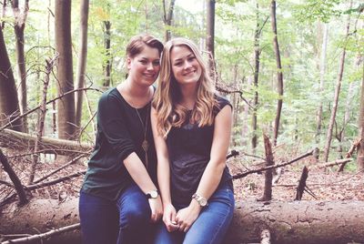 Portrait of happy female friends sitting on log in forest