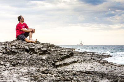 Full length of man sitting on rock at beach against sky