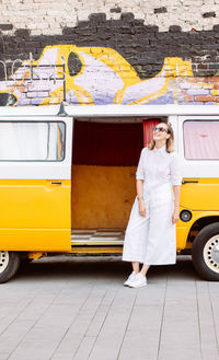 Young beautiful fashionable woman in white outfit near a yellow photo booth minibus on the street 