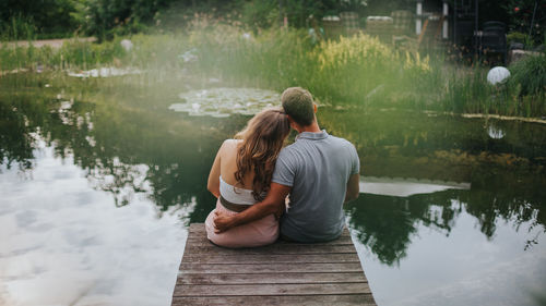 Rear view of man with pregnant wife while sitting on pier over lake