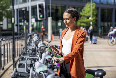 Smiling businesswoman holding mobile phone standing by electric bicycle