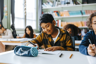 Smiling elementary schoolboy writing in book while sitting in classroom