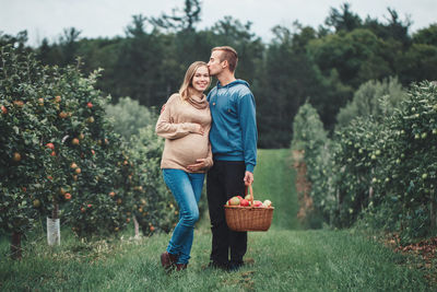 Portrait of smiling pregnant couple standing with apples in basket on grass