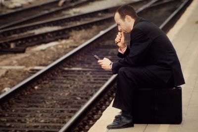 Side view of man using smart phone while sitting on briefcase at railroad station