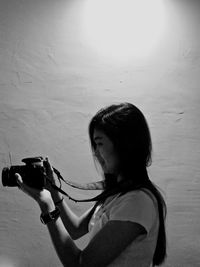 Side view of young woman photographing with camera while standing by wall at night