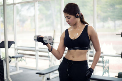 Young woman lifting dumbbell while standing against window in gym