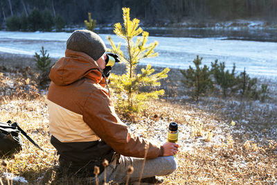 Young man in warm clothes drinking tea from thermos sitting on bank of river on winter hike