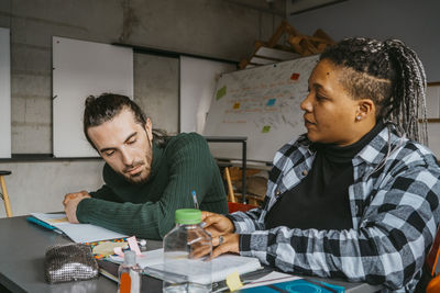 Young male university student looking at friend writing in book