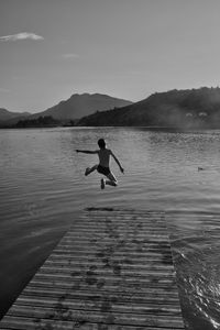 Rear view of shirtless boy jumping on pier over lake against sky