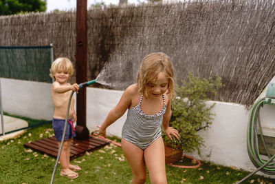 Cheerful shirtless little boy pouring water from hose on sister in swimsuit while playing together in yard on summer day