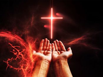 Digital composite image of hand and fire by cross against black background