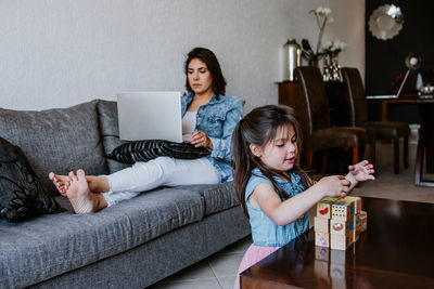 Concentrated young latin american female freelancer sitting on sofa and working remotely on laptop near cute little daughter playing with wooden cube toys at home