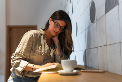 Young wistful female in casual apparel with textbook and cup of hot drink looking away in cafeteria