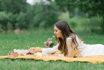 A young beautiful woman in a white dress sits on a blanket on a summer picnic on the green grass