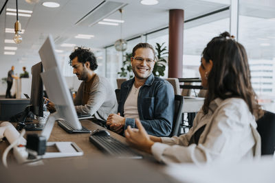 Smiling young male and female colleagues discussing while sitting by businessman at desk in office