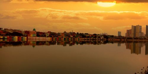 Buildings by river against sky during sunset