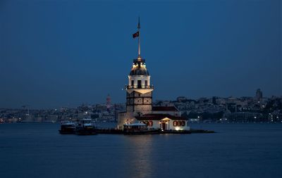 Lighthouse by sea against buildings in city against clear blue sky