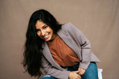 Stylish cheerful ethnic female model in trendy smart casual outfit sitting on chair and looking confidently at camera