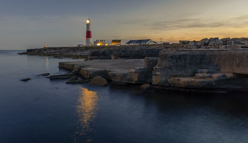 Illuminated lighthouse by sea against sky during sunset